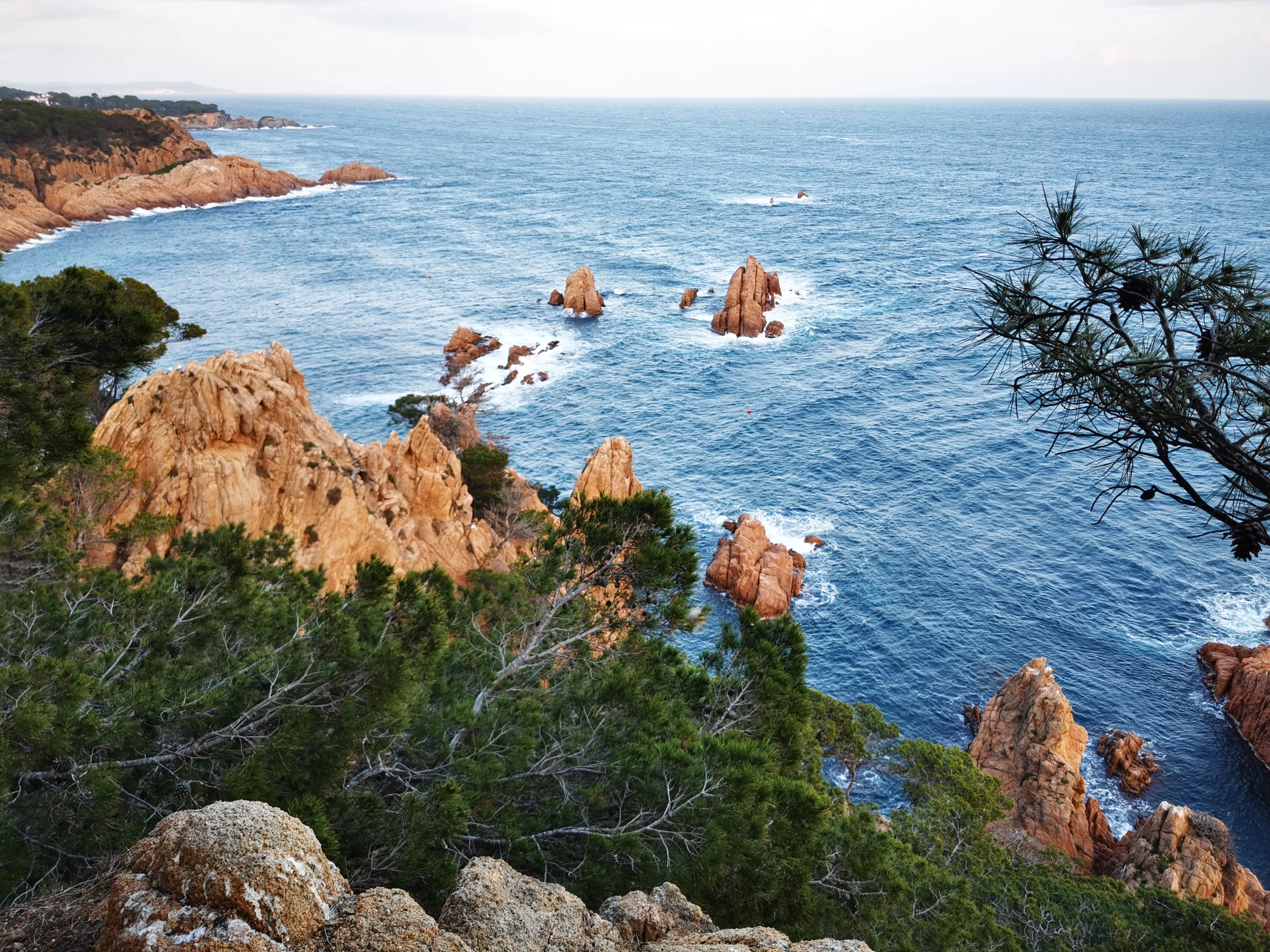 Coastal view with orange rocks, blue sea, and green trees under a partly cloudy sky.